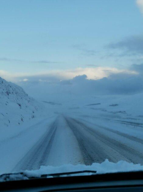 A snow-covered road stretches ahead, flanked by snow-laden fields and distant mountains under a cloudy sky. The scene is viewed from inside a vehicle.