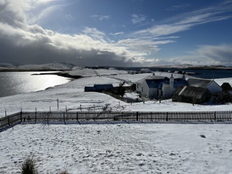 A serene, snow-covered rural landscape features a fenced property with quaint buildings near a reflective body of water under a partly cloudy sky.