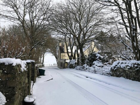 A narrow snow-covered road flanked by stone walls and leafless trees leads to a house in the distance on a winter day. Tracks from vehicles are visible in the snow.