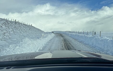 A partially cleared snowy road with a barbed wire fence and snowbanks on both sides, viewed from the interior of a vehicle.