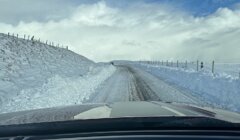 A partially cleared snowy road with a barbed wire fence and snowbanks on both sides, viewed from the interior of a vehicle.