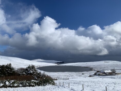 Snow-covered landscape featuring a small lake, hills, and sparse vegetation under a partly cloudy blue sky.