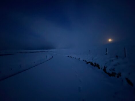 A snow-covered road illuminated by a dim light in the distance on a dark, foggy evening.