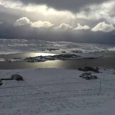 A snow-covered landscape with a body of water reflecting a cloudy sky, a small settlement is visible in the distance beneath dramatic, overcast skies.