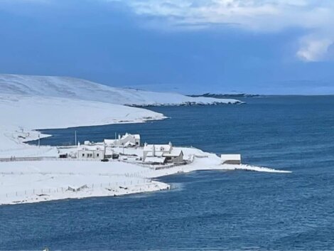 A coastal village is covered in snow beside a large body of water under a partly cloudy sky. Snow-covered hills can be seen in the background.