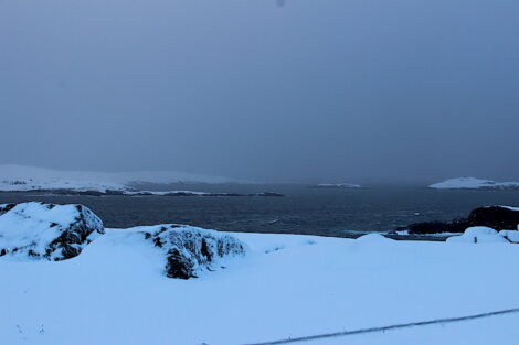 A snowy coastal landscape with rocky outcroppings, dark water, and a hazy, overcast sky in the background.