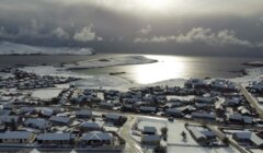 Aerial view of a snow-covered coastal town with cloudy skies and sunlight reflecting off the sea in the background.