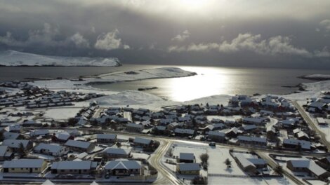 Aerial view of a snow-covered coastal town with cloudy skies and sunlight reflecting off the sea in the background.