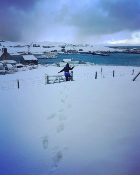 A person walks through deep snow toward a fence, with footprints visible in the snow. In the background, there is a snow-covered village with houses and a body of water.