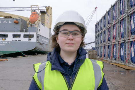 A person wearing a hard hat, safety glasses, and a high-visibility vest stands on a dock in front of a cargo ship and stacked containers.