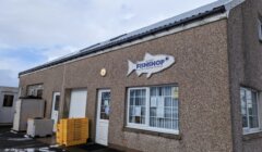 A small, beige building with a sign in the shape of a fish that reads "Fish Shop." Stacks of yellow crates are outside near the entrance. The sky above is partly cloudy.