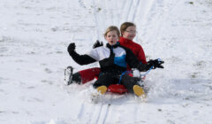 Two children wearing winter jackets and gloves are sledding down a snowy hill on a red sled. They appear to be enjoying the ride.