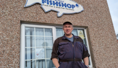 A man in a work uniform stands in front of a building with a sign shaped like a fish that reads "Blydoit Fish Shop," indicating it sells fresh fish and shellfish.