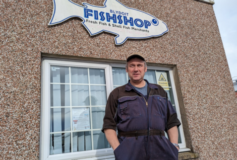 A man in a work uniform stands in front of a building with a sign shaped like a fish that reads "Blydoit Fish Shop," indicating it sells fresh fish and shellfish.