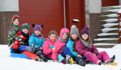 A group of children dressed in winter clothing sit together on the snow, smiling and enjoying the snowy day.