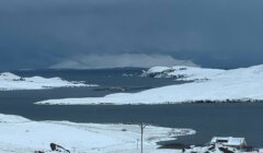 Snow-covered landscape with a body of water and small islands in the foreground. A dark, cloudy sky looms above the distant mountains.