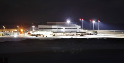 A modern airport terminal at night is illuminated with bright lights, surrounded by snow-covered grounds and several parked service vehicles and aircraft.