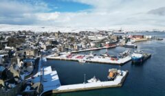 Aerial view of a coastal town with a snow-covered landscape, small boats in multiple docks, and a partially cloudy sky overhead.