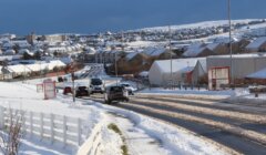 Snow-covered suburban street with several vehicles driving, surrounded by houses, and a mix of cloudy and blue sky in the background.