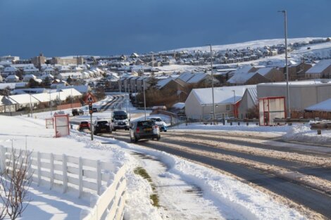 Snow-covered suburban street with several vehicles driving, surrounded by houses, and a mix of cloudy and blue sky in the background.