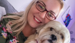 A woman with blonde hair and glasses smiles while holding a small, fluffy dog. Colorful artwork is visible in the background.