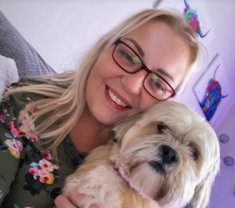 A woman with blonde hair and glasses smiles while holding a small, fluffy dog. Colorful artwork is visible in the background.