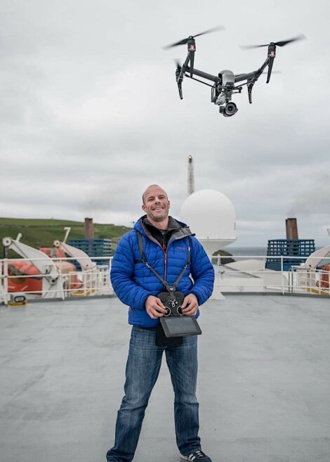 A man in a blue jacket controls a drone using a remote controller on a ship deck with lifebuoys and other equipment in the background.