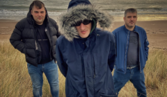 Three men in winter jackets stand on a grassy dune with a beach and ocean in the background. The man in the center wears a hooded coat with fur trim.