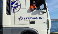 A person wearing a safety vest is seated in the driver's seat of a white Streamline truck parked near water. The truck features the company logo and website.