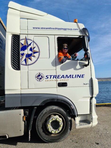 A person wearing a safety vest is seated in the driver's seat of a white Streamline truck parked near water. The truck features the company logo and website.