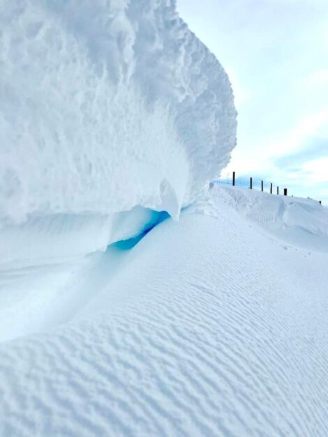 A close-up view of a large snowdrift with a blue tint beneath an overhanging edge. Snow-covered landscape with a fence in the background.