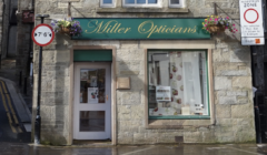 A storefront with a green sign reading "Miller Opticians." Above the door and windows are hanging flower baskets. A nearby sign indicates a pedestrian zone with restricted vehicle access.