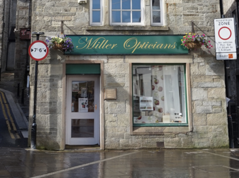 A storefront with a green sign reading "Miller Opticians." Above the door and windows are hanging flower baskets. A nearby sign indicates a pedestrian zone with restricted vehicle access.