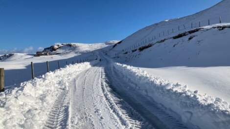 A snow-covered road winds through a hilly landscape with tire tracks visible, flanked by snow walls. A clear blue sky stretches above.