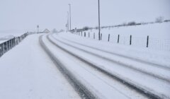 A snow-covered road with visible tire tracks, flanked by fences and streetlights, stretches into the distance under a cloudy sky. Road signs can be seen near the road's curve.