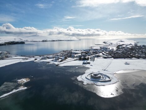 Aerial view of a coastal town surrounded by snow and ice, with a calm body of water and a snow-covered island in the background under a partly cloudy sky.