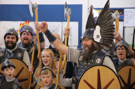 A group of people dressed in medieval costumes, some holding shields and axes, are gathered together, with one person raising their fist in the air. Artwork is visible on the wall in the background.