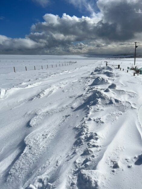 A snow-covered road with snow drifts on both sides under a cloudy sky. Power lines run along the right side of the image.