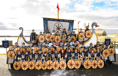 A large group of people dressed as Vikings stand in front of a Viking ship replica, holding shields and wearing traditional costumes. A body of water and cloudy sky are visible in the background.