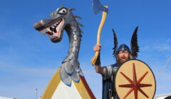 A man in Viking attire stands next to a dragon-headed structure. He holds a shield and raises an axe while wearing a helmet with black wings. The sky is clear and blue.