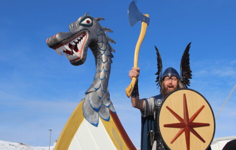 A man in Viking attire stands next to a dragon-headed structure. He holds a shield and raises an axe while wearing a helmet with black wings. The sky is clear and blue.