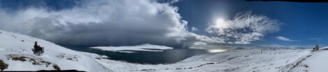 A panoramic view of a snowy landscape by the sea, with partly cloudy skies and visible sun. An individual sits in a wheelchair on the left side of the image, and a small building is seen on the right.