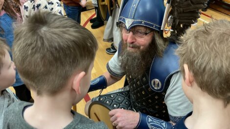 A person dressed in Viking attire, complete with helmet and armor, interacts with a group of children in an indoor setting.