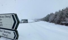 Snow-covered road with a vehicle driving in the distance. Road signs pointing to Whalsay and Out Skerries ferry directions are visible in the foreground. Snowfall obscures visibility.