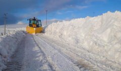 A snowplow clears a path through a thick layer of snow, with snow banks piled high on both sides of the road under a blue sky with clouds.