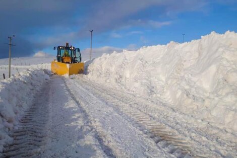 A snowplow clears a path through a thick layer of snow, with snow banks piled high on both sides of the road under a blue sky with clouds.