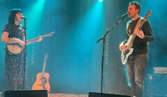 Two musicians performing on stage; a woman singing and playing a banjo, and a man singing and playing an electric bass guitar. A guitar is placed on a stand between them, with blue spotlights overhead.