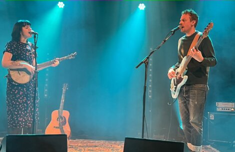 Two musicians performing on stage; a woman singing and playing a banjo, and a man singing and playing an electric bass guitar. A guitar is placed on a stand between them, with blue spotlights overhead.