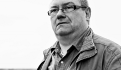 Black-and-white photo of a man with glasses wearing a jacket and a collared shirt, looking off into the distance against a light background.