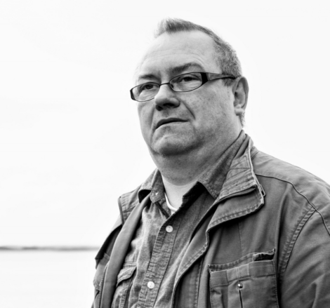 Black-and-white photo of a man with glasses wearing a jacket and a collared shirt, looking off into the distance against a light background.